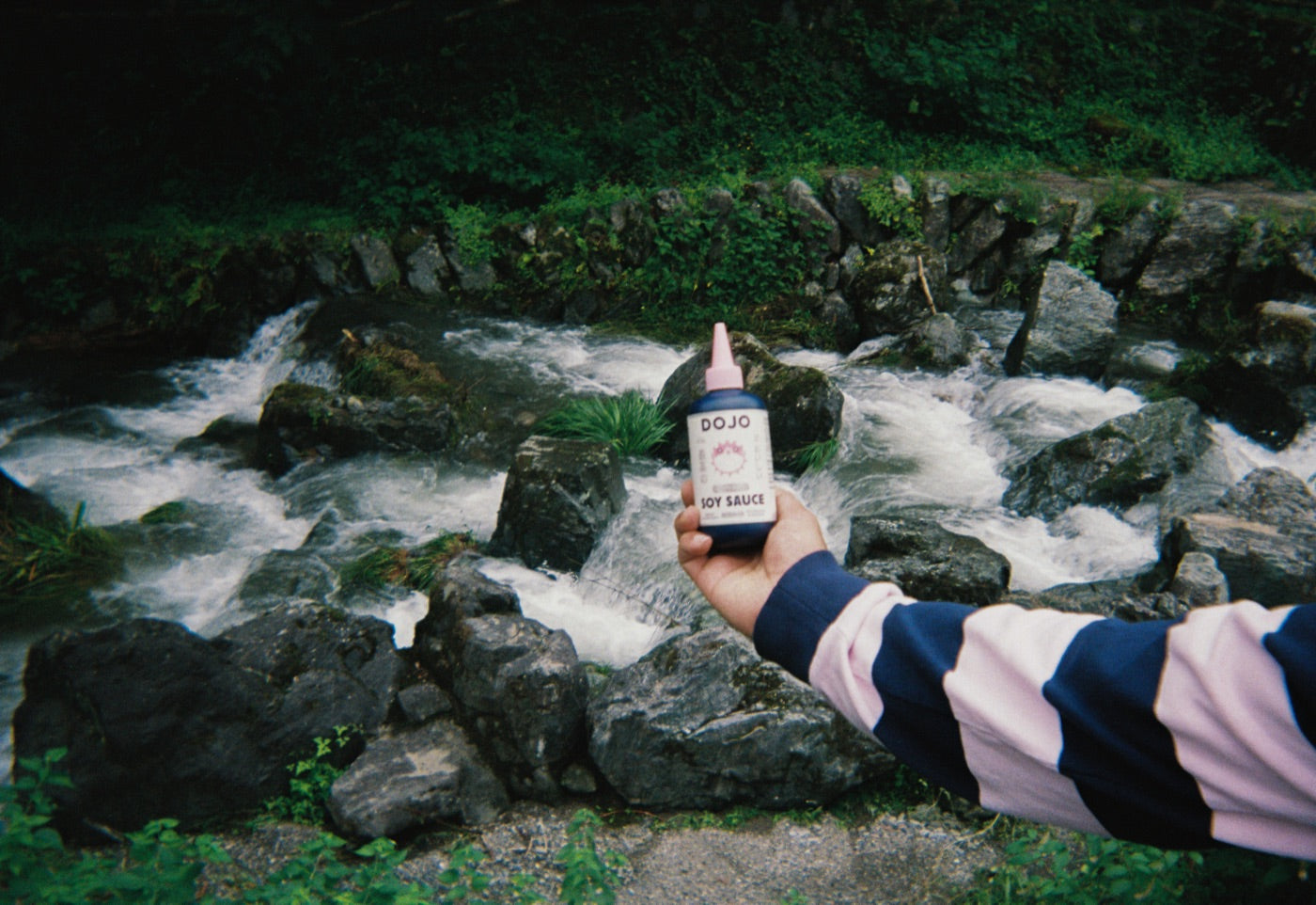 A hand holding the Dojo bottle in front of a rushing mountain stream