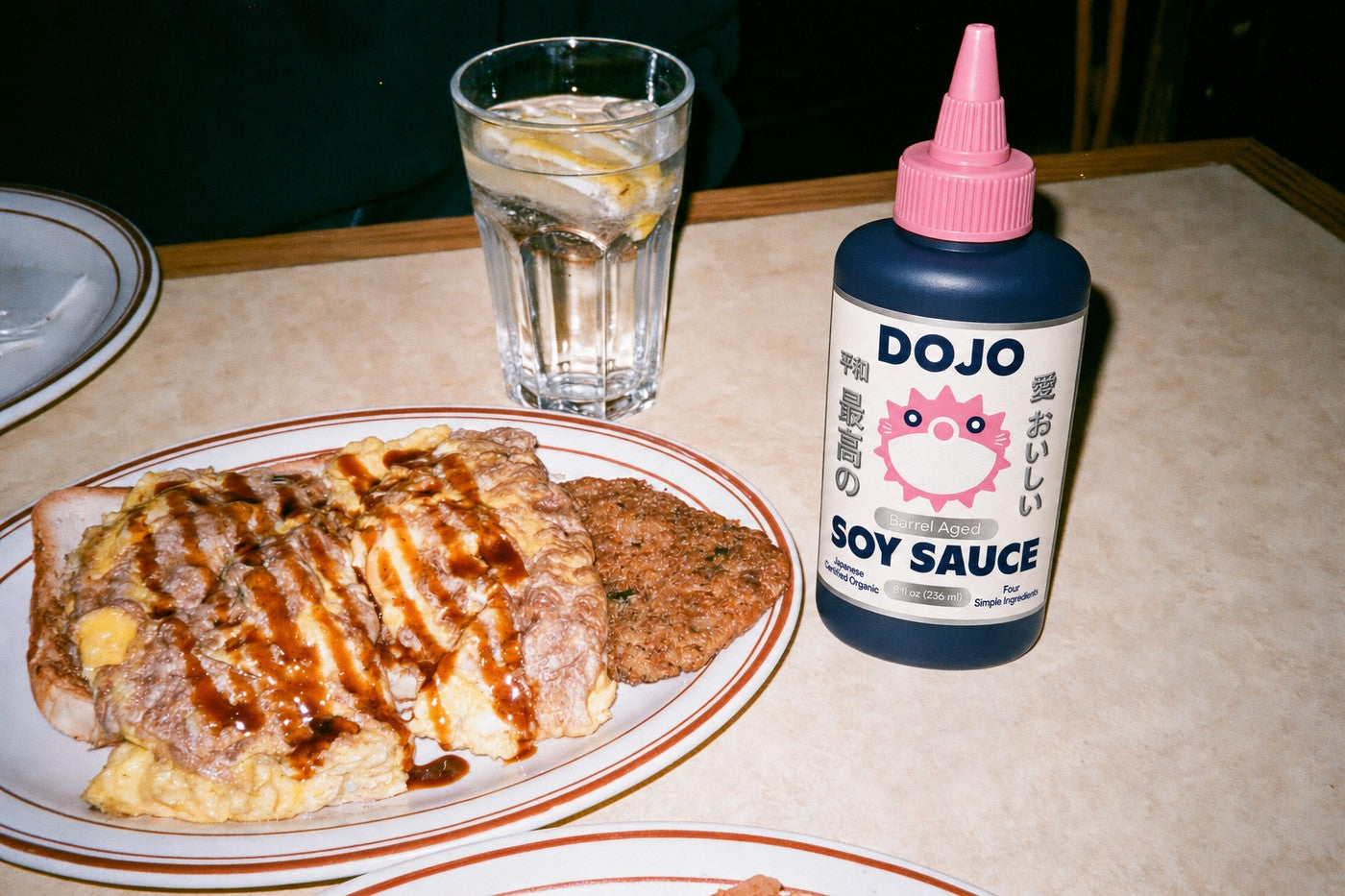 The Dojo bottle standing beside a breakfast sandwich with eggs and hash browns, drizzled with soy sauce