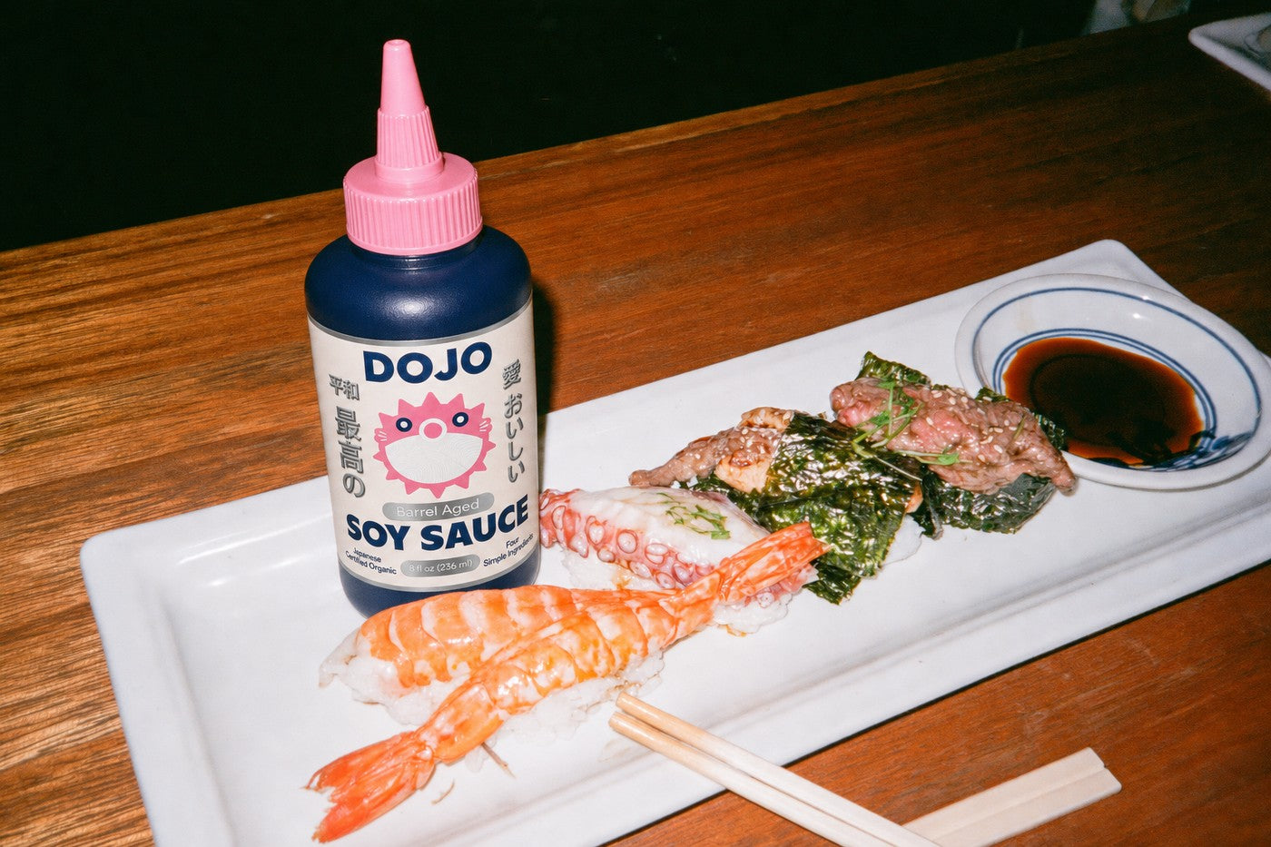 The Dojo bottle standing on a sushi counter beside a plate of shrimp nigiri and a dish of soy sauce