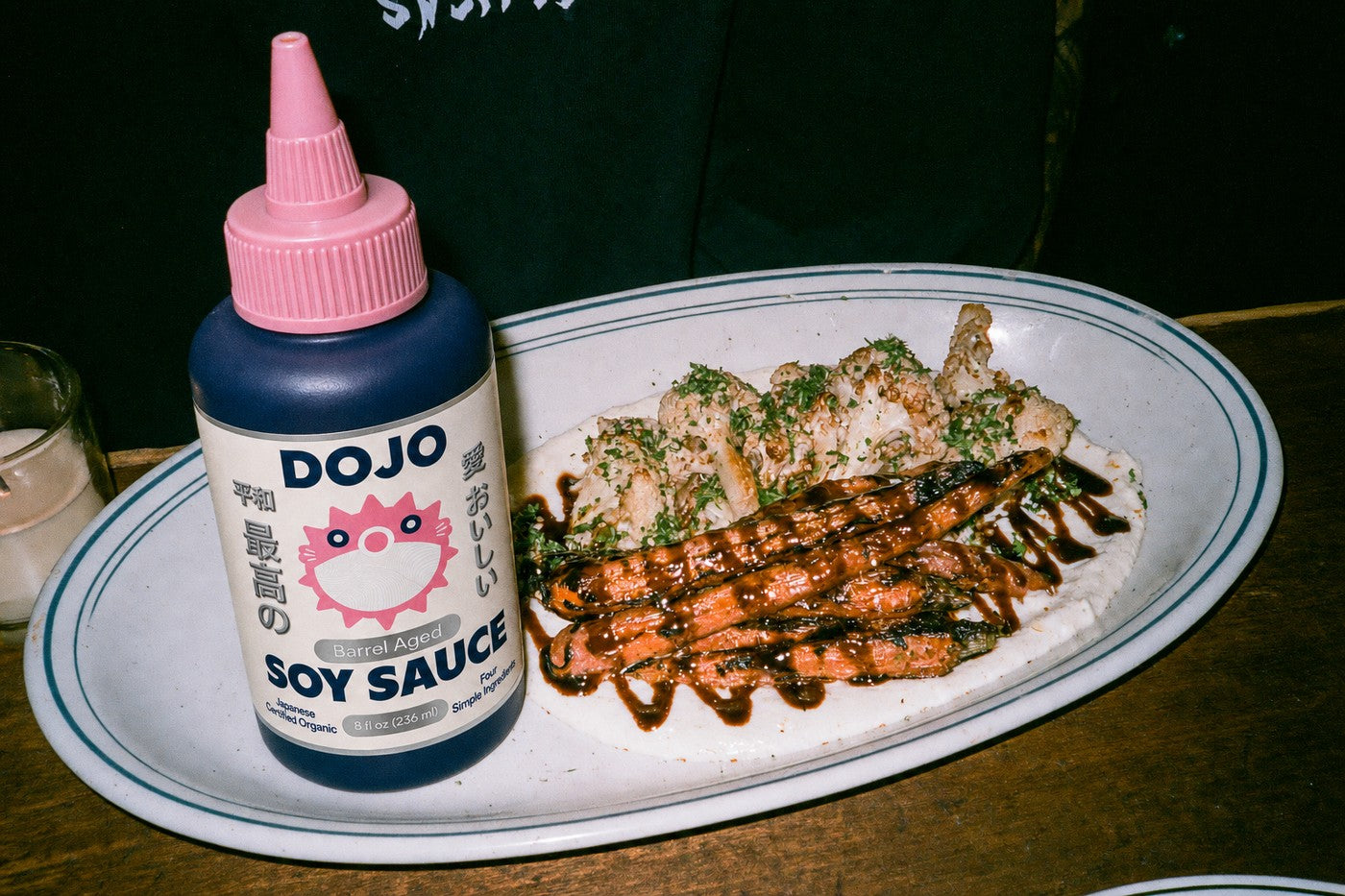 The Dojo bottle standing beside a platter of charred carrots and cauliflower over labneh, finished with soy sauce
